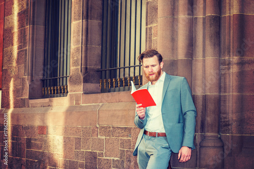 Foto American Graduate Student with beard, mustache studying in New York, wearing cadet blue suit, standing against wall with windows on campus, reading red book, thinking