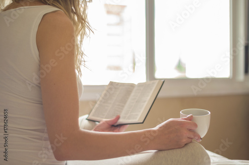Jovem mulher caucasiana lora lendo livro, bíblia, bebendo café, chá na cama com raios de sol pela janela, manhã cedo. photo