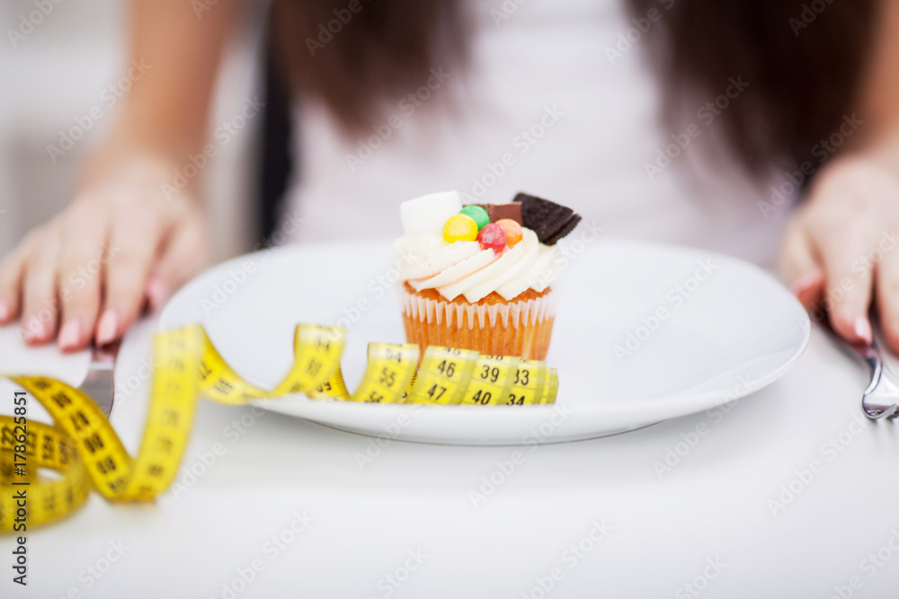 Young dieting woman sitting in front of plate with delicious cream tart cakes, looking at forbidden food with unhappy and hungry expression, studio Young dieting woman sitting in front of plate with delicious cream tart cakes, looking at forbidden food with unhappy and hungry expression, studio