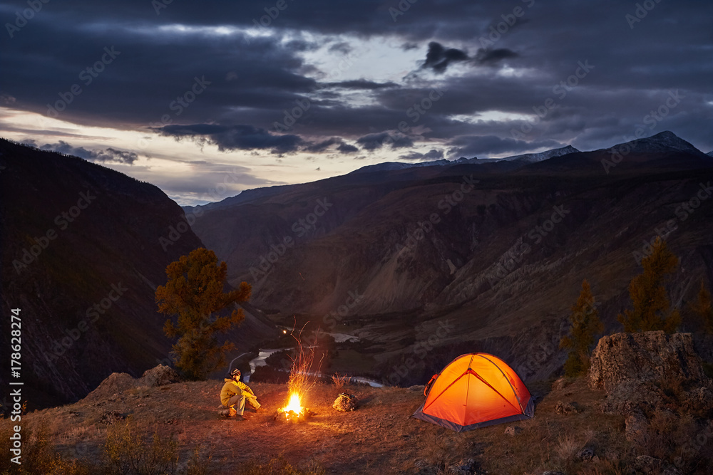 A man near illuminated tent and campfire in mountains in dawn