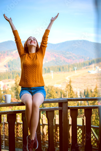 Cute and nice young woman drinking coffee at terrace on a sunny morning in mountain. Pretty lady in stylish casual yellow sweater and shorts. Warm soft cozy image, breakfast on the balcony 