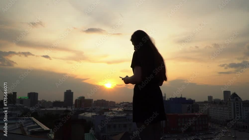 The girl looks in the smartphone against the background of a yellow decline. Young girl using the smartphone sitting on the roof.. The girl looks in the smartphone against the background of a yellow decline. Young girl using the smartphone sitting on the roof..