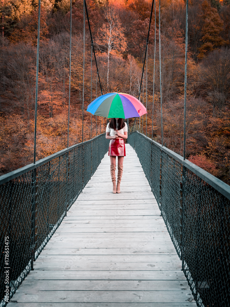 Beautiful Caucasian European girl with a red skirt, high boots and white top on a bridge holding a colorful rainbow umbrella