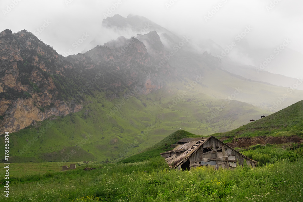 old wooden house in the thick green grass on a background of mountains in the fog