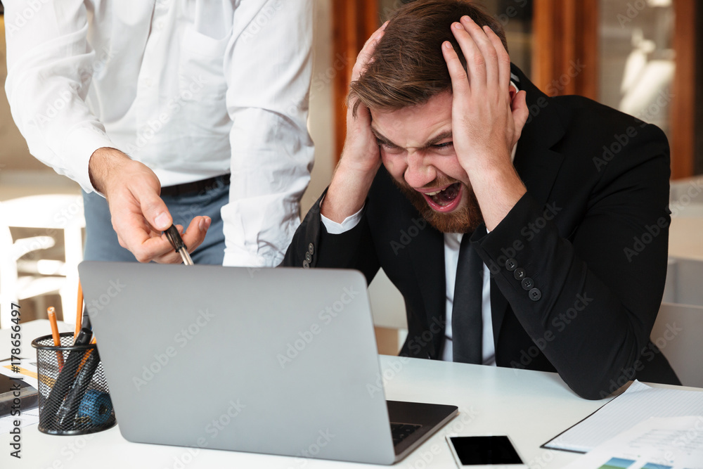 Young angry bearded businessman in formalwear holding his head, looking at computer while his partner pointing with pen on laptop screen in office Young angry bearded businessman in formalwear holding his head, looking at computer while his partner pointing with pen on laptop screen in office
