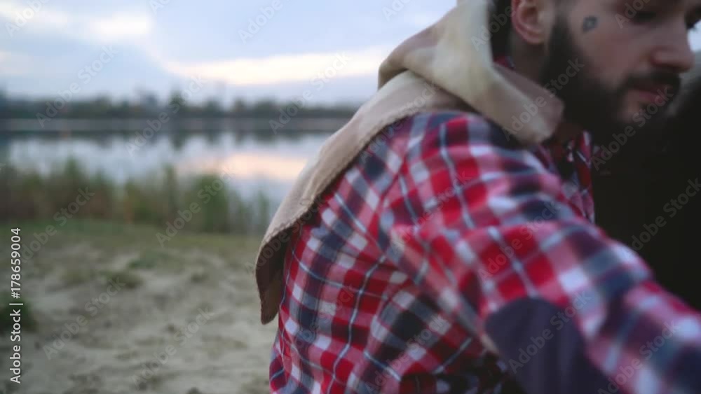 Couple in love sitting around the bonfire at the lake, talking, drinking hot tea and fry marshmallows Couple in love sitting around the bonfire at the lake, talking, drinking hot tea and fry marshmallows