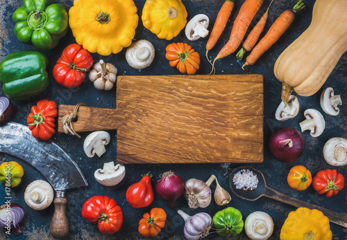 Tomatoes, onions, mushrooms, carrots, pumpkin, patissons, garlic, spices and knives on dark blue grunge plywood background with wooden chopping board in center. Top view, copy space