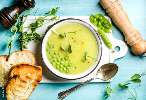 Light summer green pea cream soup in bowl with sprouts, bread toasts and spices. White ceramic board in the center, turquoise blue wooden background. Top view
