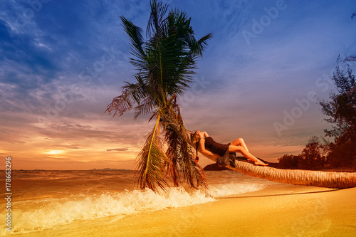 Young beautiful woman in black dress and the palm tree above sea on sunset time. Tropical vacation concept.