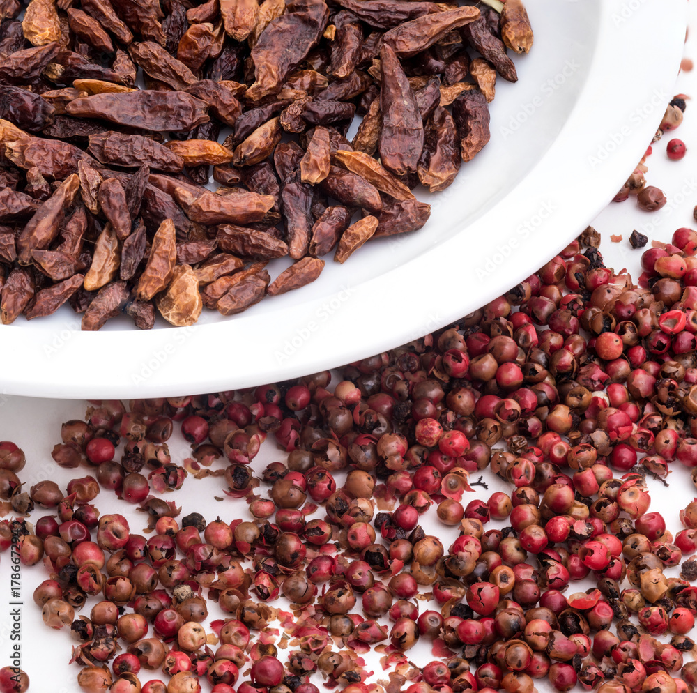 Dried small red peppers in close-up on a white plate and pink pepper grains on a white table. Dried small red peppers in close-up on a white plate and pink pepper grains on a white table.