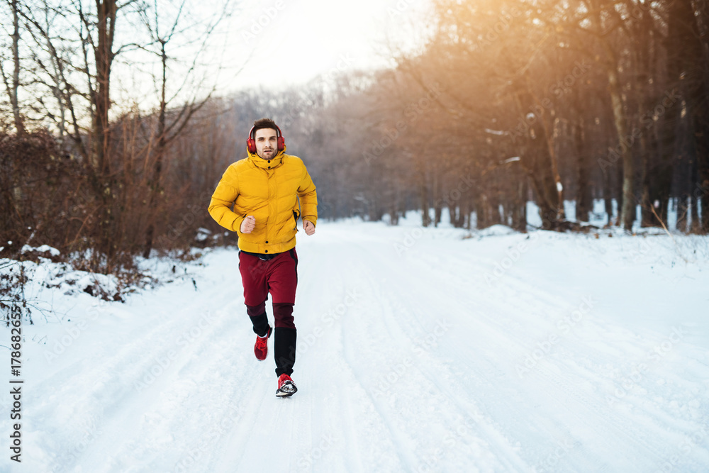 Fit young man in winter sportswear with headphones running on snow covered winter road in the morning through the forest.