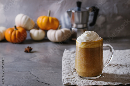 Glass of spicy pumpkin latte with whipped cream and cinnamon standing on gray kitchen table with coffeepot and decorative white pumpkins. Coffee beans and spices above. Atmospheric breakfast
