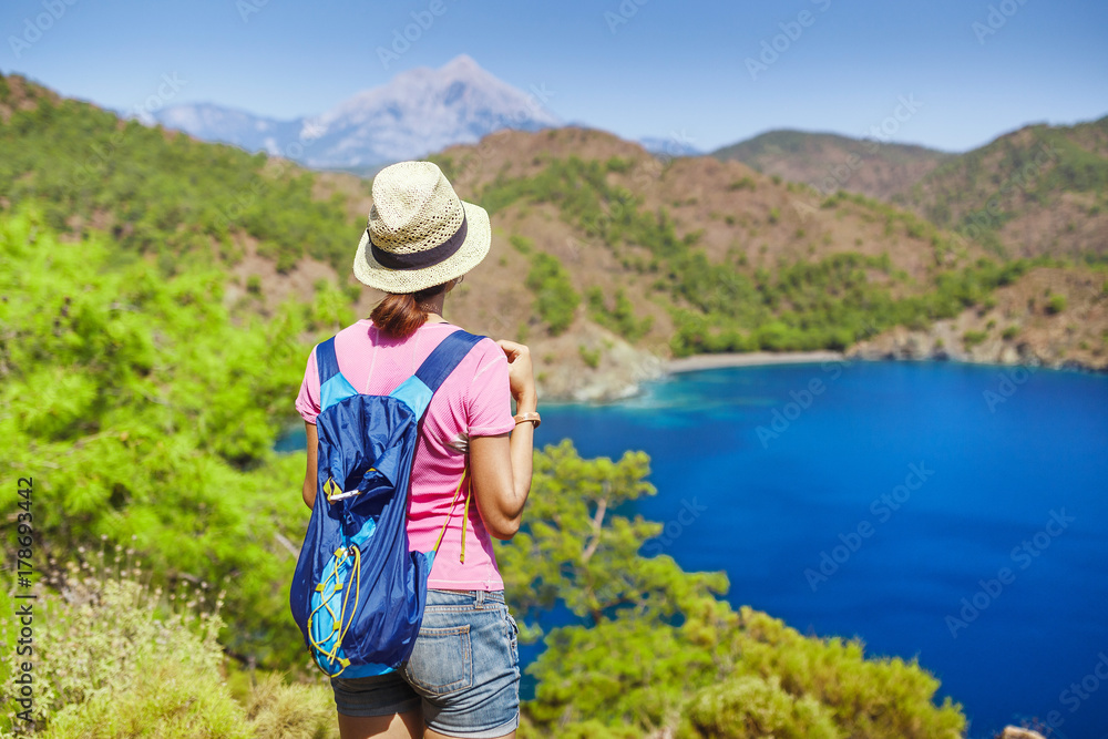 Young woman traveler with backpack standing on cliff's edge and looking to a mediterranean sea landscape, summer vacation concept