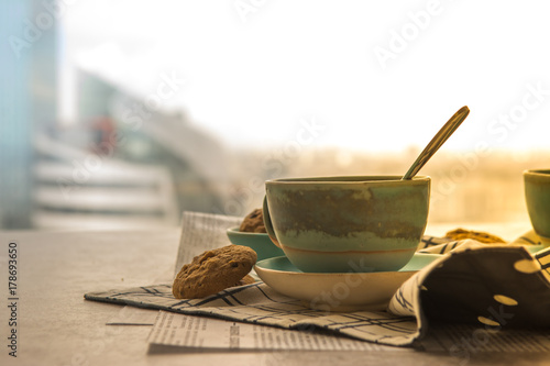 Cup of black coffee with spoon on supported dish on fabric on black and white newspaper is put on hard table in the early morning with bright yellow light and some biscuits.