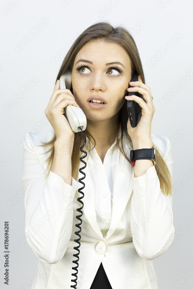 The beautiful girl holds two phones. Beautiful girl in a business suit on a white background with two phones. Different emotions on the face when talking on the phone The beautiful girl holds two phones. Beautiful girl in a business suit on a white background with two phones. Different emotions on the face when talking on the phone