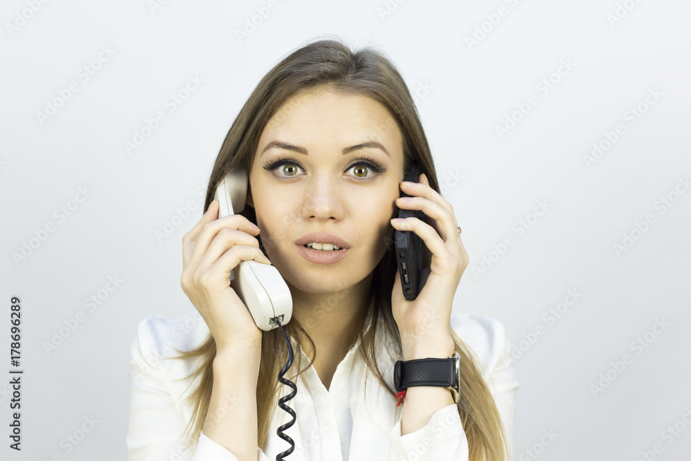 The beautiful girl holds two phones. Beautiful girl in a business suit on a white background with two phones. Different emotions on the face when talking on the phone The beautiful girl holds two phones. Beautiful girl in a business suit on a white background with two phones. Different emotions on the face when talking on the phone