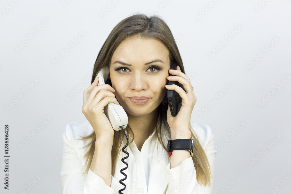 The beautiful girl holds two phones. Beautiful girl in a business suit on a white background with two phones. Different emotions on the face when talking on the phone The beautiful girl holds two phones. Beautiful girl in a business suit on a white background with two phones. Different emotions on the face when talking on the phone