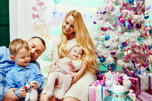 Beautiful family with children in warm sweaters poses before a wall and rich Christmas tree