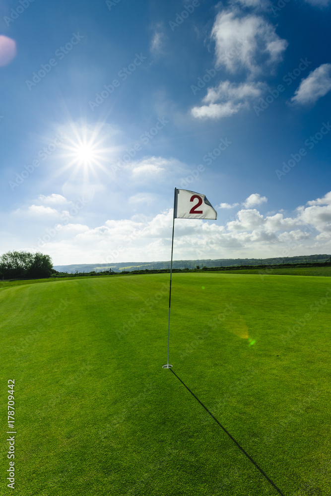 Obraz premium View of a green golf course, hole and flag on a bright sunny day. Sport, relax, recreation and leisure concept. Summer landscape with sunbeams