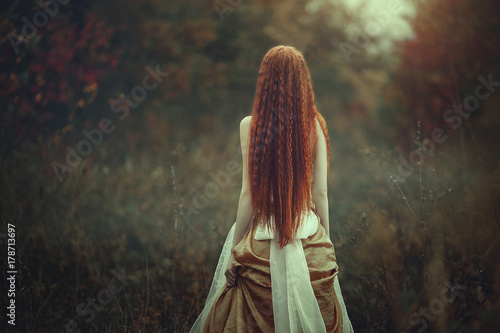 Fototapeta A beautiful young woman with very long red hair as a witch walks through the autumn forest