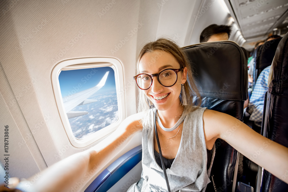 Fototapeta premium Young happy woman making selfie photo sitting on the aircraft seat near the window during the flight in the airplane