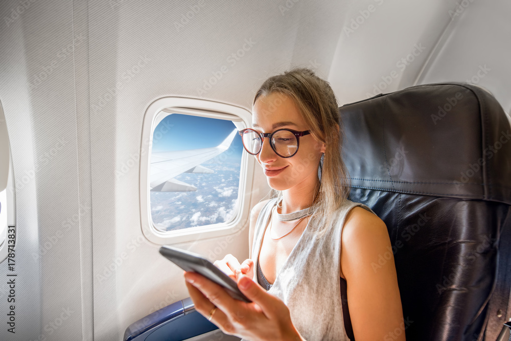 Fototapeta premium Young woman sitting with phone on the aircraft seat near the window during the flight in the airplane