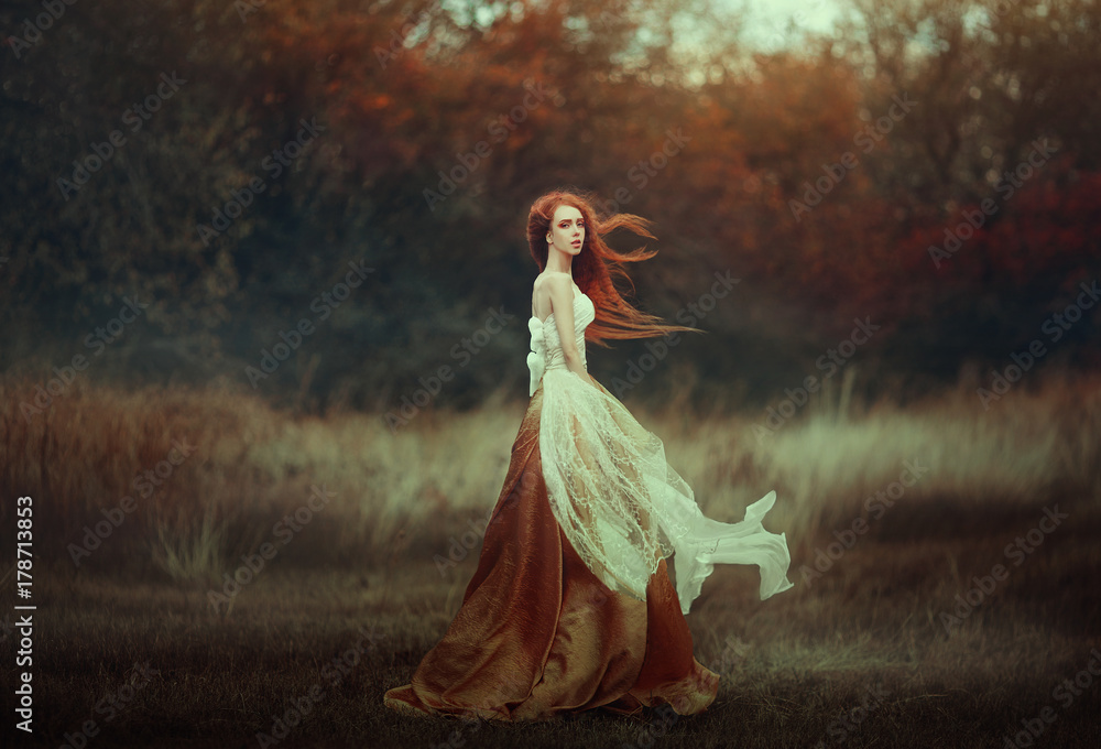 Beautiful young woman with very long red hair in a golden medieval dress walking through the autumn forest. Long red hair develops in the wind.