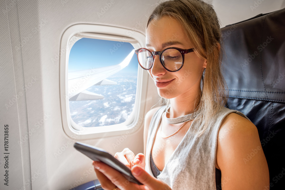 Fototapeta premium Young woman sitting with phone on the aircraft seat near the window during the flight in the airplane