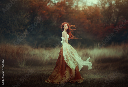 Obraz na plátně Beautiful young woman with very long red hair in a golden medieval dress walking through the autumn forest