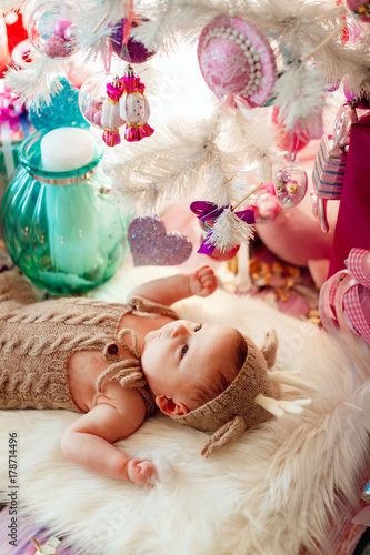 Baby in deer suit lies on soft white pillow under white Christmas tree with pink toys