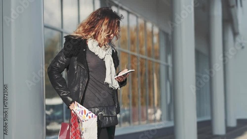 Woman in sun glasses a black leather jacket, black jeans with shopping bags walking and talking on mobile phone in front of mirrored windows on the Street