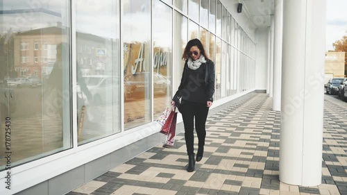 Woman in sun glasses a black leather jacket, black jeans with shopping bags, enjoying the walk in front of a mirrored store window on the street. Steadicam shot
