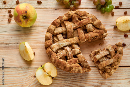 Homemade american apple pie piece and fresh fruits - apples and grapes on brown wooden table background. Homemade classical fruit tart with raisins. Top view