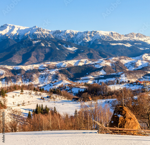 winter landscape in Romania with Haystacks, traditional village, snowy Bucegi mountains (Carpathians) in Transylvania. Romanian nature, countryside landscape, scenery in wintertime. 