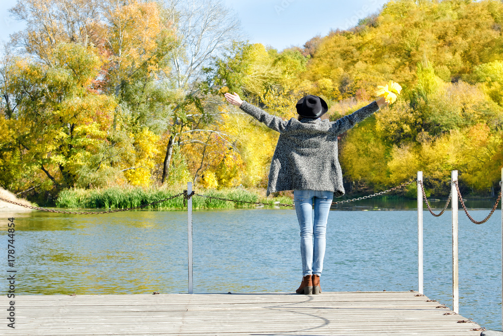 Girl in the hat is standing on the dock with her hands up. Autumn, sunny. Back view