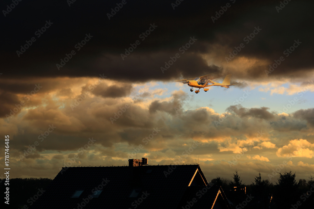Samolot awionetka w burzowych chmurach nad dachami budynków. - obrazy, fototapety, plakaty 