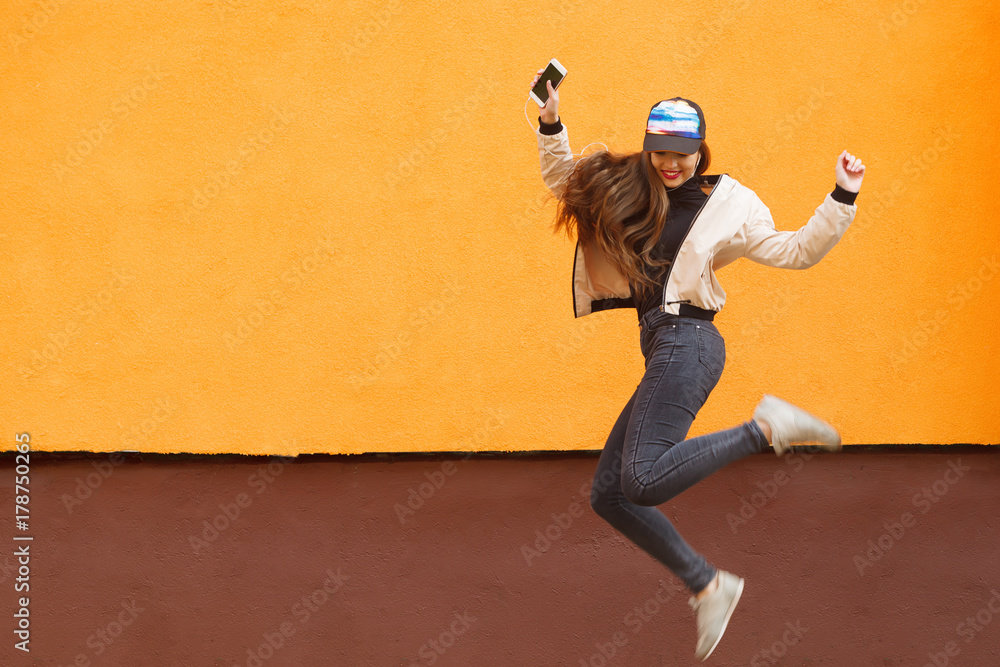 Young hipster girl jumping against the orange and listened to music by headphones by smartphone. Urban style.