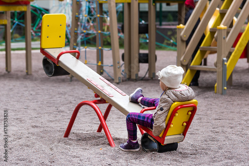 Quadro su tela Little cute caucasian girl in jacket and hat sitting alone on a seesaw swing at a playground outdoors