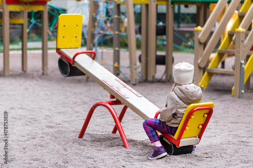 Foto Little cute caucasian girl in jacket and hat sitting alone on a seesaw swing at a playground outdoors