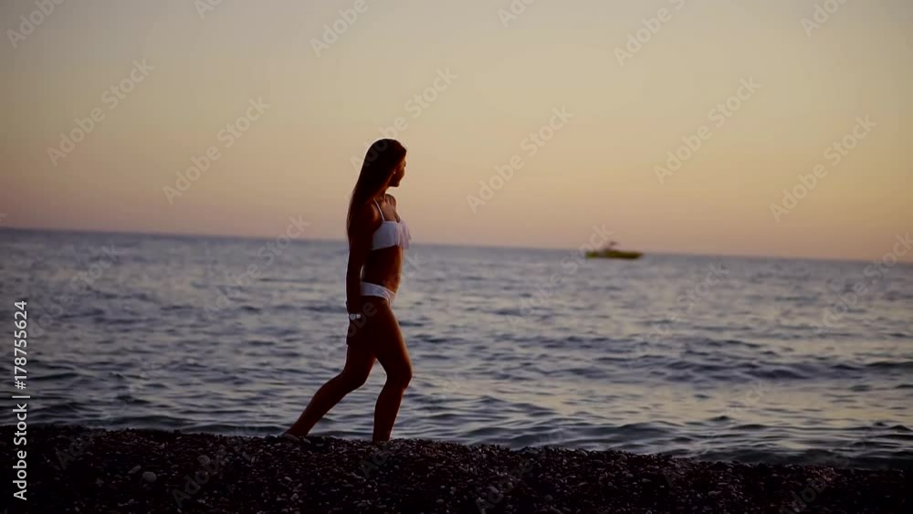A young woman is walking along the beach in the evening, the waves are beating against the shore, in the distance there is a boat A young woman is walking along the beach in the evening, the waves are beating against the shore, in the distance there is a boat