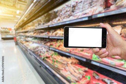 Hand holding smartphone with white blank screen and blurred supermarket ham sausage background