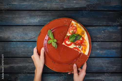 Woman's hands holding a plate with one pieces of pizza margarita with tomato slices, olives and basil leaves. Top view