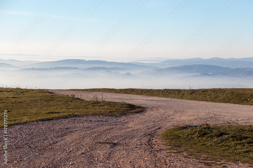 A mountain road pointing toward some distant mountains and hills in the midst of fog and mist