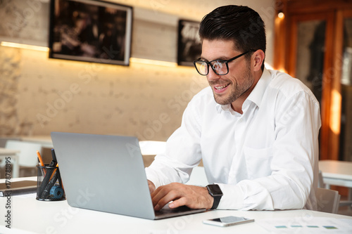 Close-up photo of handsome smiling businessman in white shirt using laptop in office