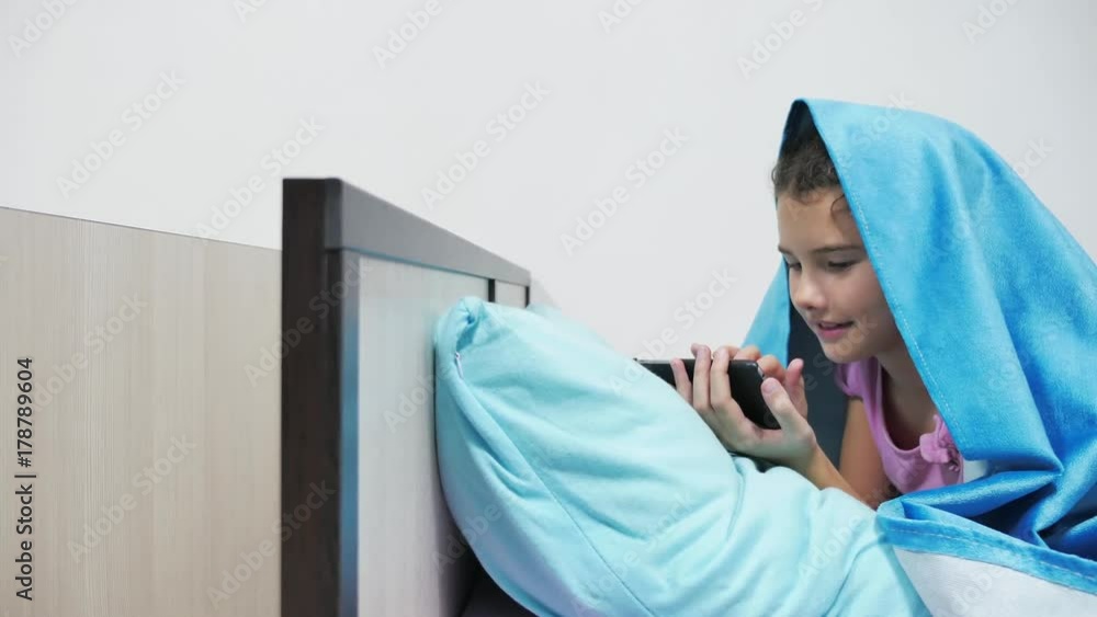 girl teenager under a blanket playing in a smartphone. little girl before bed lies in bed surfing indoor on the internet girl teenager under a blanket playing in a smartphone. little girl before bed lies in bed surfing indoor on the internet