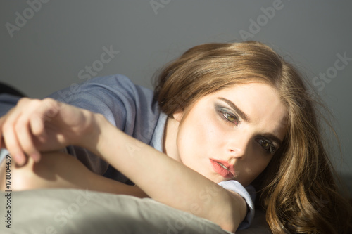 Indoors portrait of attractive young woman wearing blue shirt and lying on the bed. Looking away. Long curly hair. Grey background