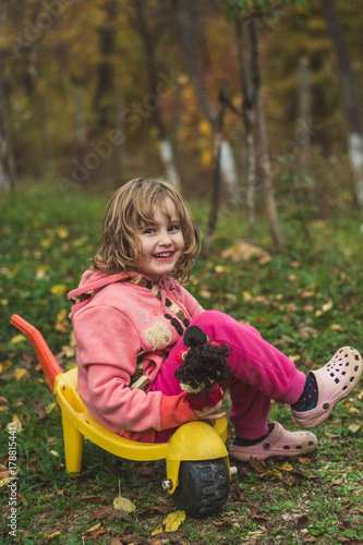 Young girl laughing and sitting in a yellow wheelbarrow with her doll in a hand.