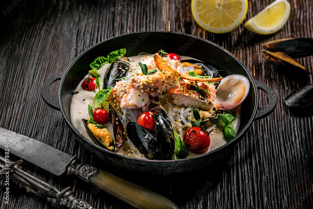 Seafood meal made of sea bass fish, mussels, crab meat and cockles with vegetables and rice noodles in delicious cream sauce in a black bowl. Seafood meal made of sea bass fish, mussels, crab meat and cockles with vegetables and rice noodles in delicious cream sauce in a black bowl.