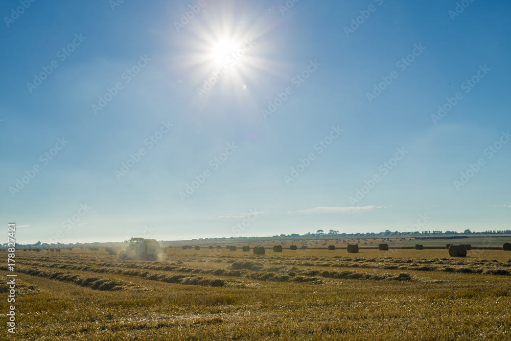 Yellow wheat field with straw bales after harvesting on a sunny day in Normandy, France. Country landscape with sunbeams in the blue sky, agricultural fields in summer. Industrial agriculture concept.