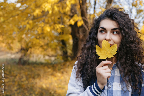 Happy curly woman holding autumn leaf in hand and posing in autumn park.